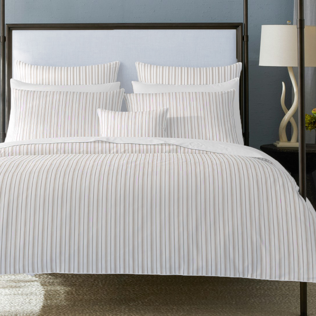 A neatly made bed with a white duvet cover and matching striped pillows, set in a bedroom with a dark metal canopy bed frame, blue-gray walls, and bedside tables with lamps.